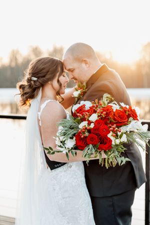 Romantic wedding couple sharing intimate moments at sunset with vibrant red rose bouquet between them.
