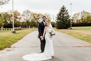 A couple in formal wedding attire shares a romantic moment on a tree-lined path during an autumn day.