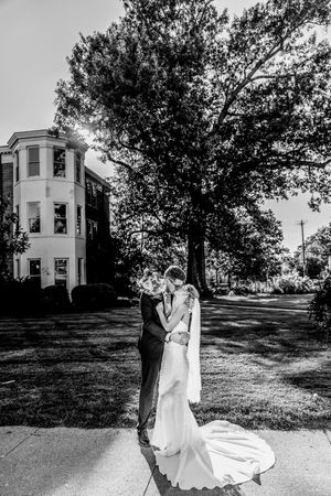 Black and white photo of a bride and groom embracing outdoors near a historic building and large trees.