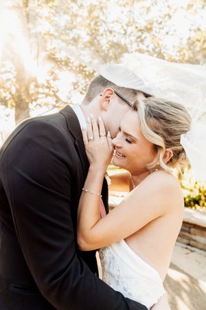 Bride and groom share a romantic moment under a flowing veil in golden sunlight, bride smiling as groom leans in.