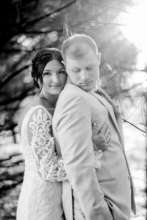 A couple in wedding attire embraces in a dreamy black and white outdoor setting with soft sunlight filtering through trees.