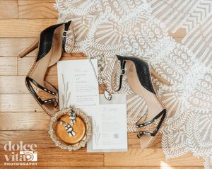 Wedding details including black stiletto heels, invitations and lace backdrop on wooden floor, flat lay photography.