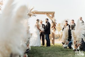 An outdoor wedding ceremony under a wooden arch decorated with pampas grass and dried florals.