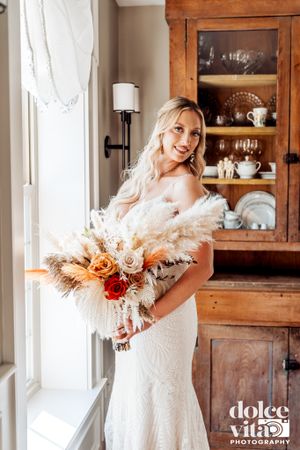 A bride in white gown holds a vibrant bouquet with roses and pampas grass near a vintage wooden cabinet.