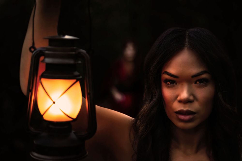A beautiful and mysterious woman holds up a lantern in the dark. In the background something blurry lurks.