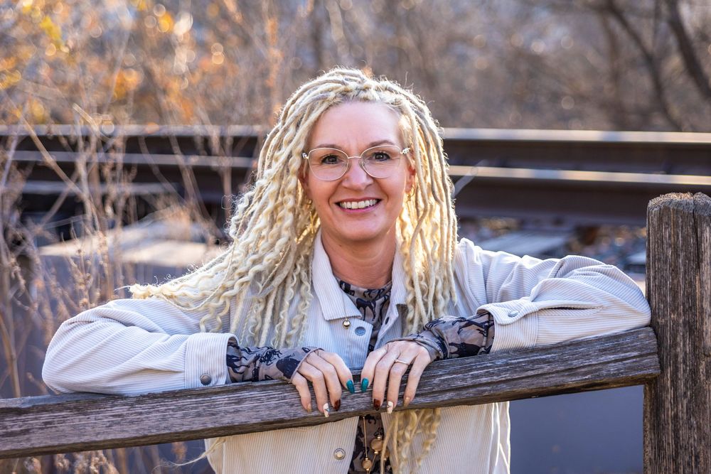 A woman with bright blonde dreads poses on an old wooden fence near train tracks.