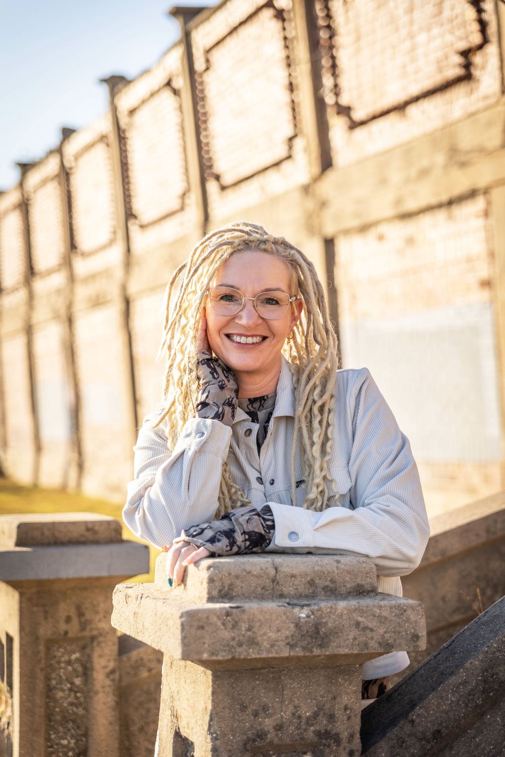 A woman with bright blonde dreads poses near a concrete wall.