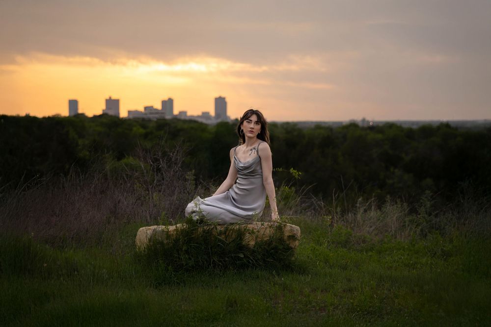 A woman in a silver dress poses on a rock during sunset with the Fort Worth skyline in the background.