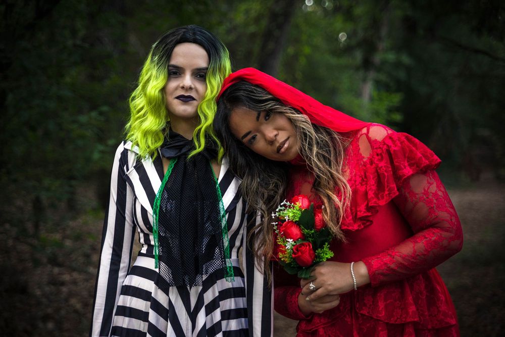 A woman in a red dress with a red veil leans her head on the shoulder of another woman wearing black and white stripes and green hair.