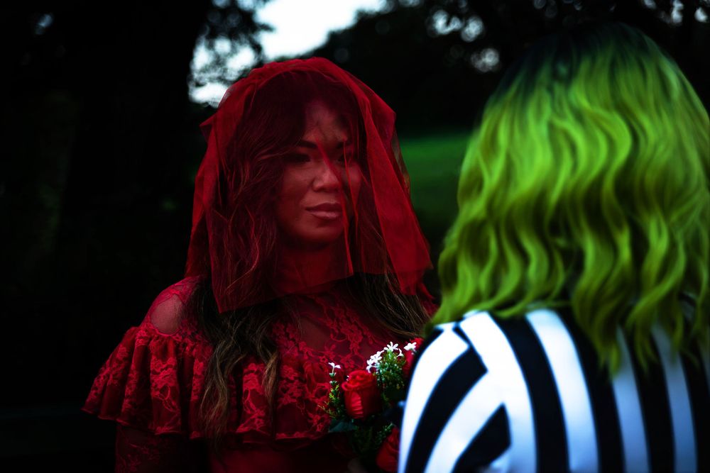 A woman wearing a red dress and red veil looking at another woman wearing black and white stripes and green hair.