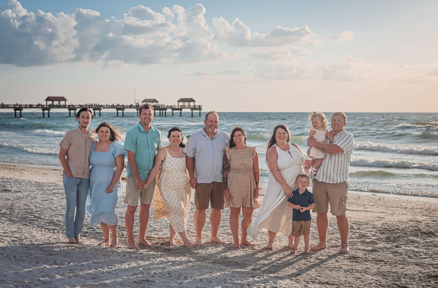 Extended family poses together on a sunny beach at sunset with waves and pier in background. Extended family photography