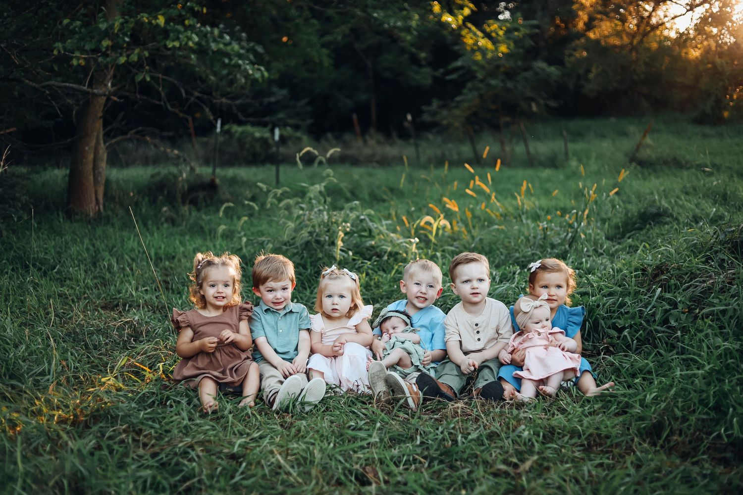 Group of children sitting together outdoors in autumn with greenery and trees in background.