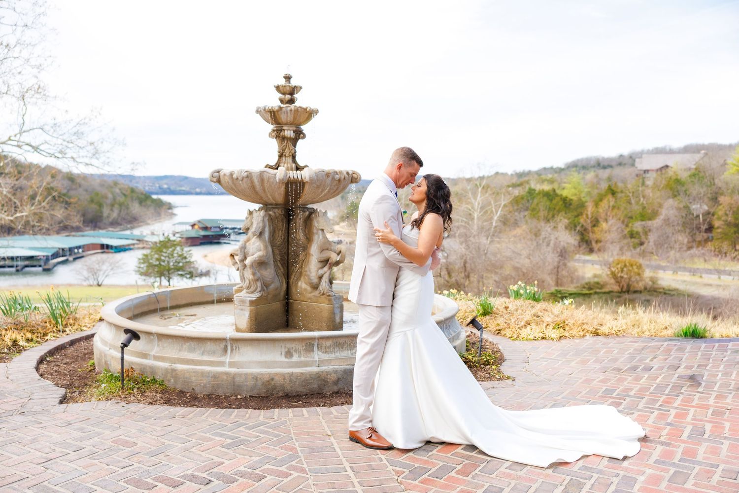 Bride and groom share a romantic kiss beside an ornate stone fountain with a scenic lake view at their outdoor wedding at Big Cedar Lodge.
