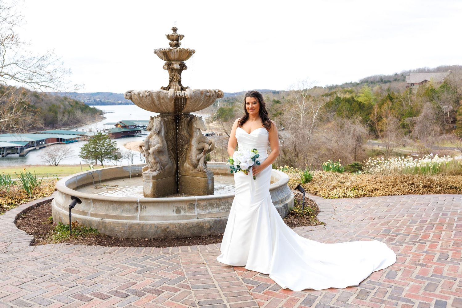 Bride in white mermaid gown holding bouquet poses by stone fountain with lake view backdrop at Big Cedar Lodge.