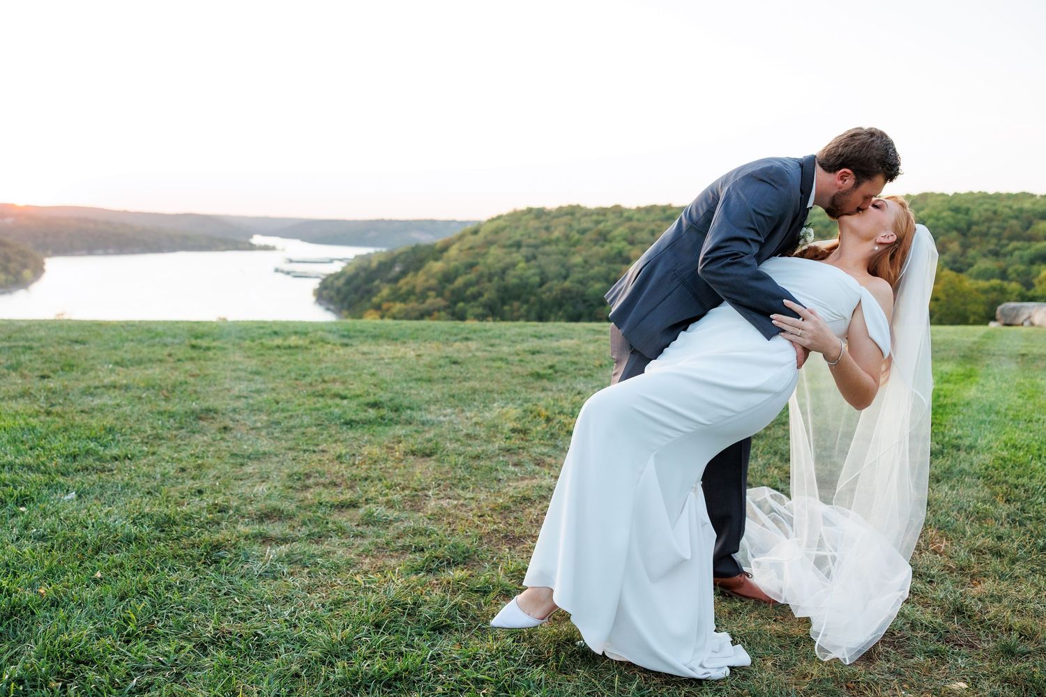 Groom dips bride in white gown for romantic kiss on green hillside overlooking scenic river at Big Cedar Lodge.