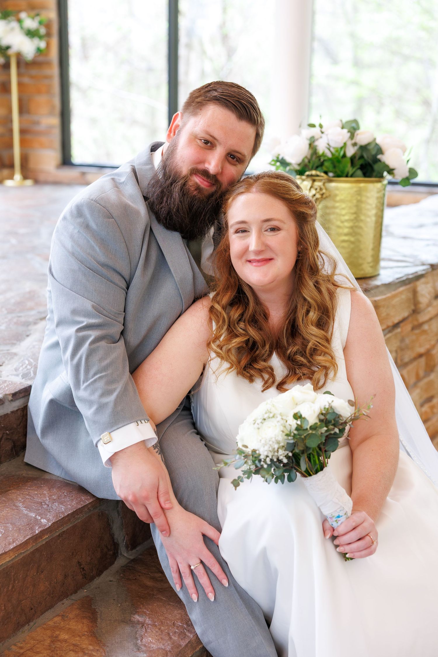 Bride and groom sitting together smiling, she holds white bouquet, he wears gray suit, indoor wedding portrait at Stonegate Glass Chapel.