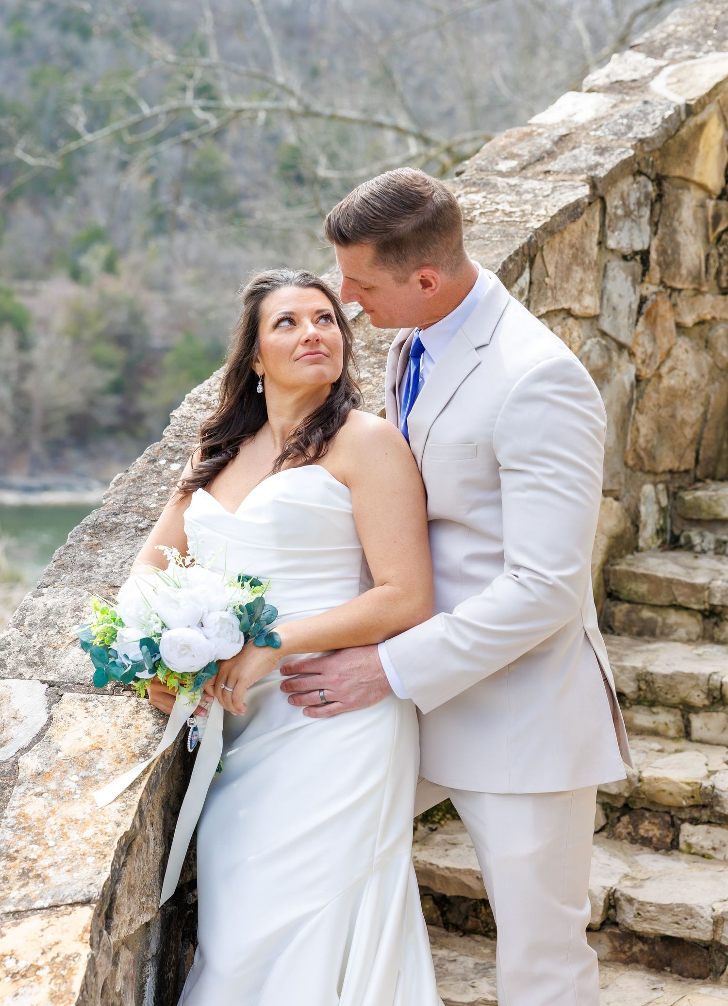 Bride and groom pose romantically by a stone wall outdoors, she holds white roses bouquet, he wears a tan suit with blue tie.