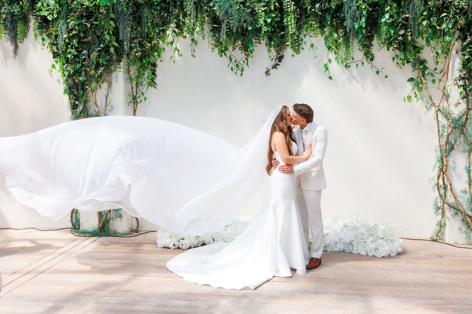 Bride and groom sharing a romantic kiss under lush greenery arch with flowing white veil billowing in the breeze at Greenhouse Two Rivers.