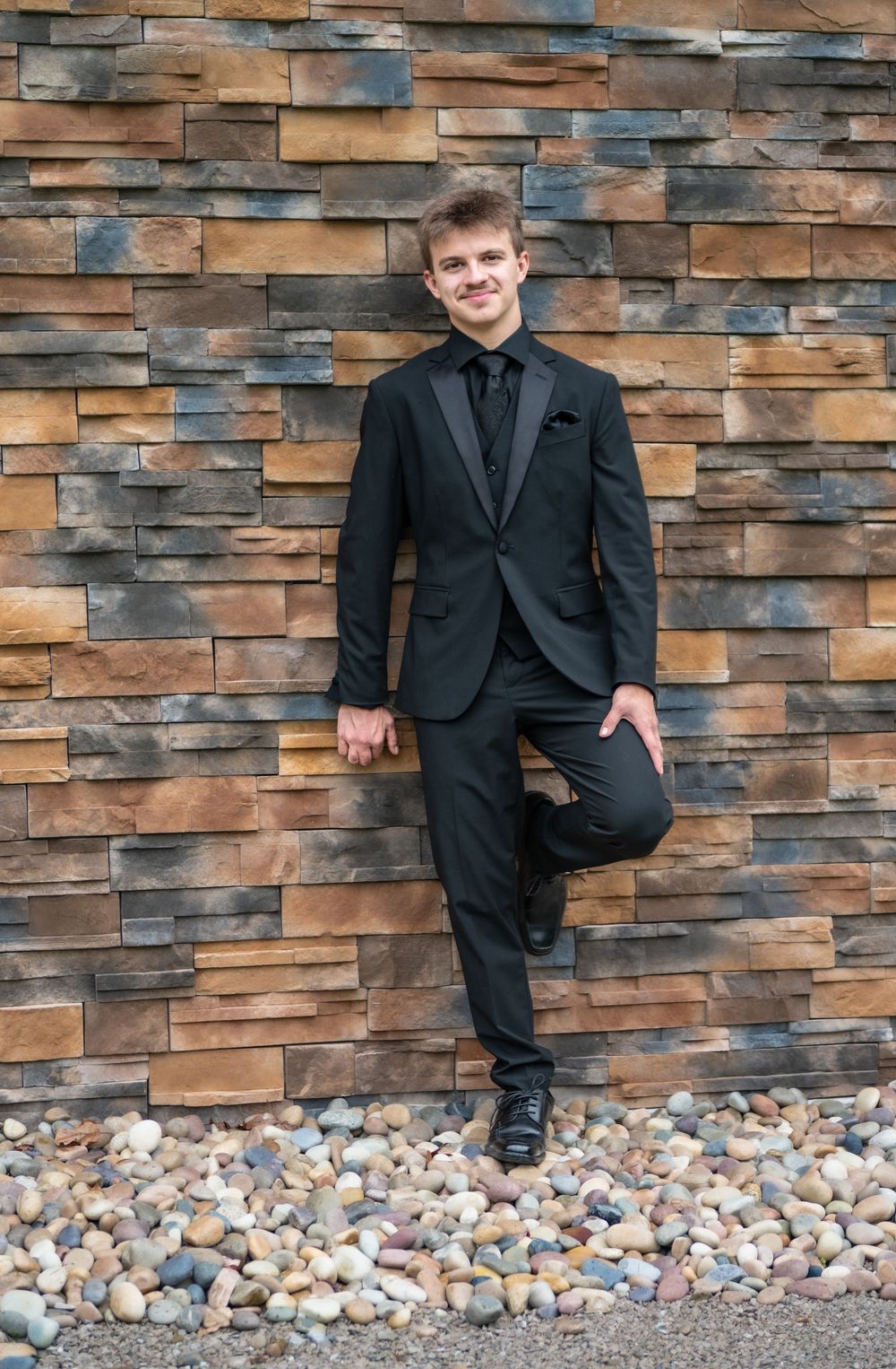 A person in a black suit poses against a textured stone wall background.