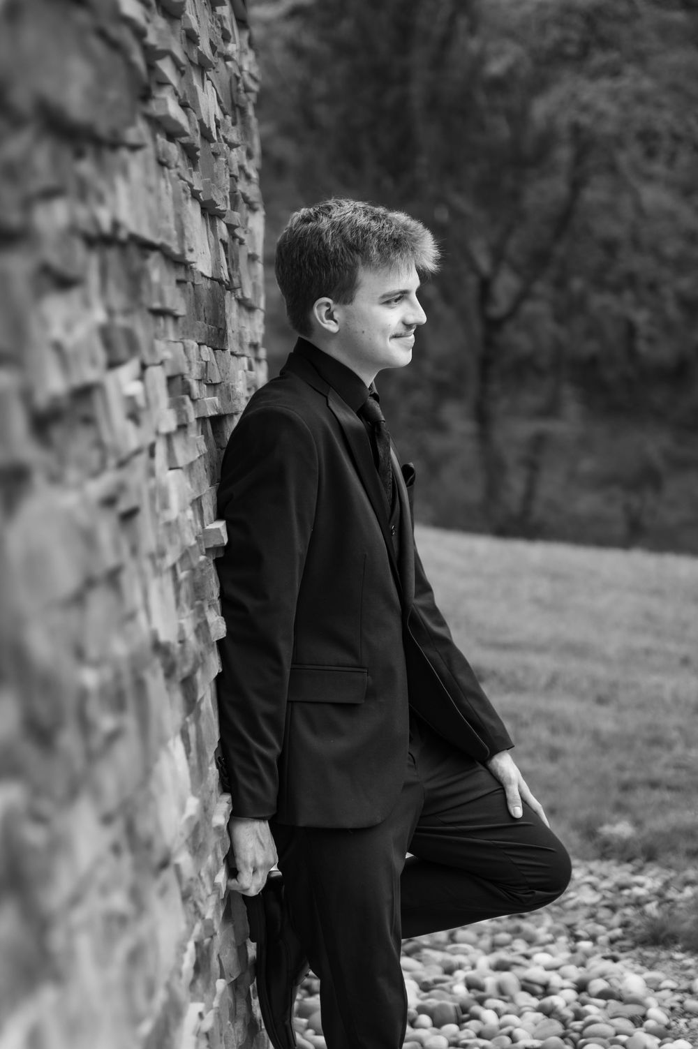 Black and white portrait of a person in formal attire leaning against a stone wall.