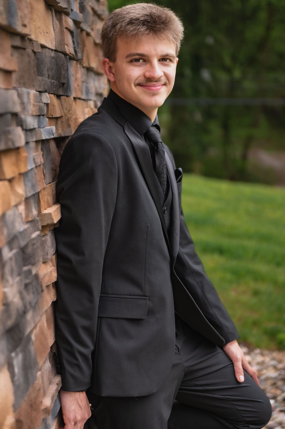 A person in formal attire smiles while posing against a decorative stone wall outdoors.