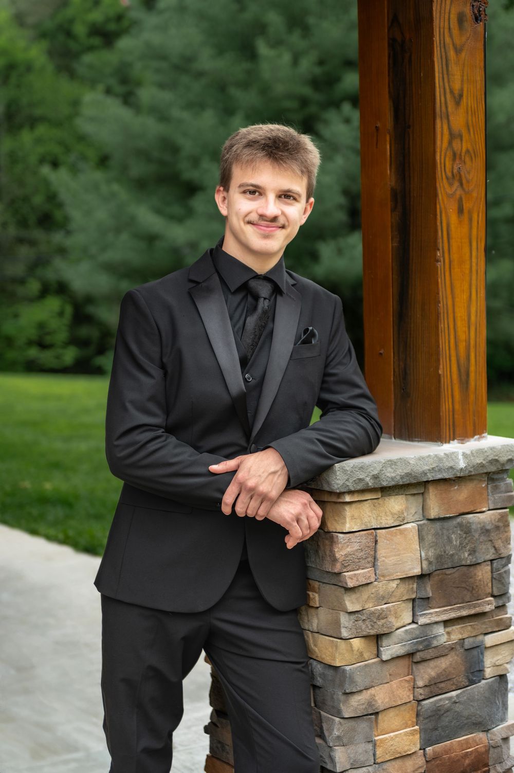 A person in a black tuxedo poses by a stone pillar with greenery in the background.