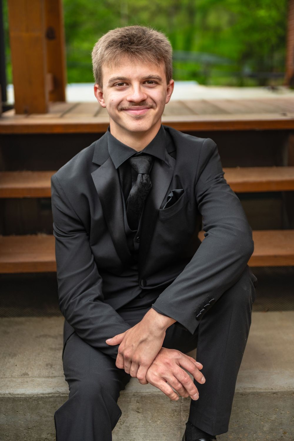 A person in a black suit sits on wooden steps with a warm smile.