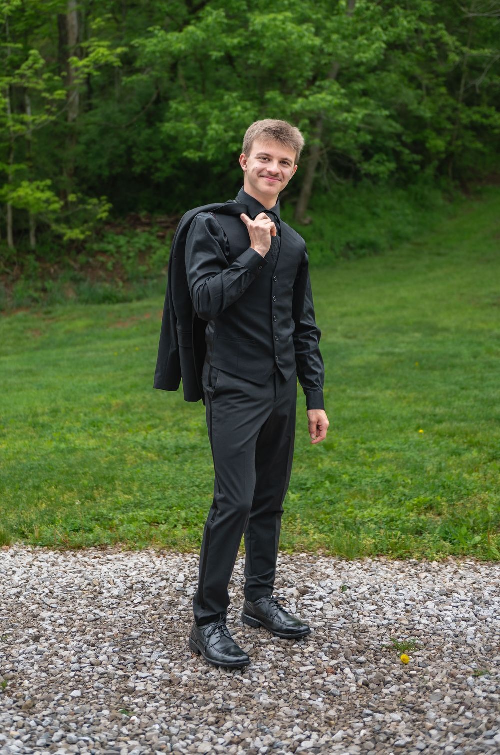 A person in a black suit carries their jacket over their shoulder on a gravel path.