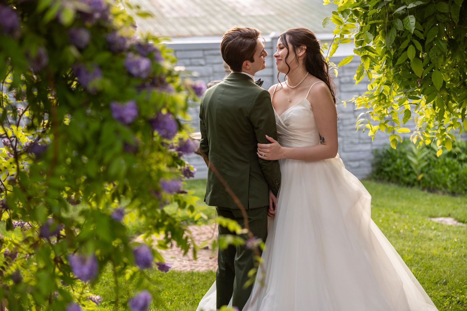 A bride and groom share a romantic moment surrounded by lush green foliage and purple flowers.