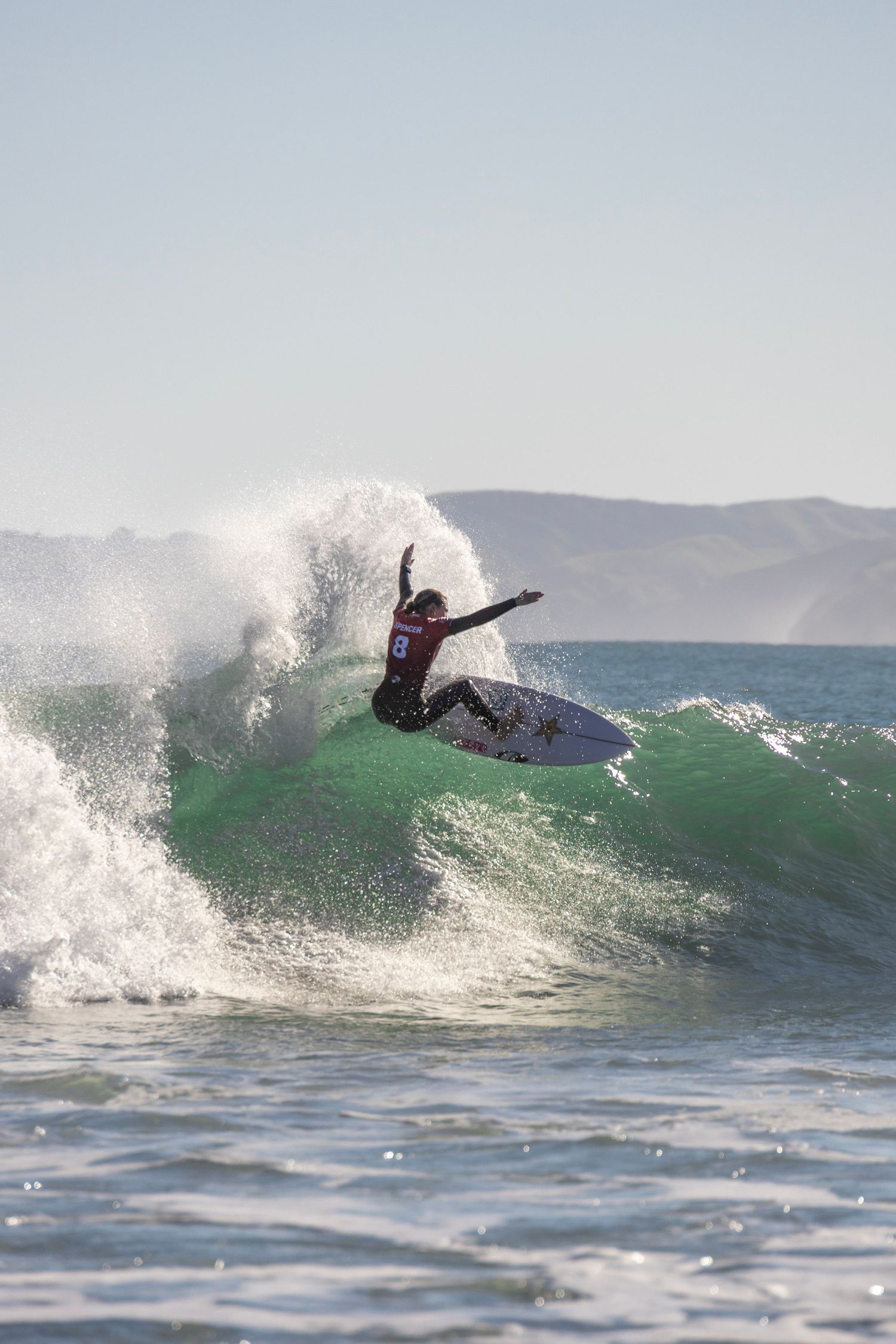 A surfer rides a clean green wave under soft morning light, with gentle ripples around them and distant land visible on the horizon.