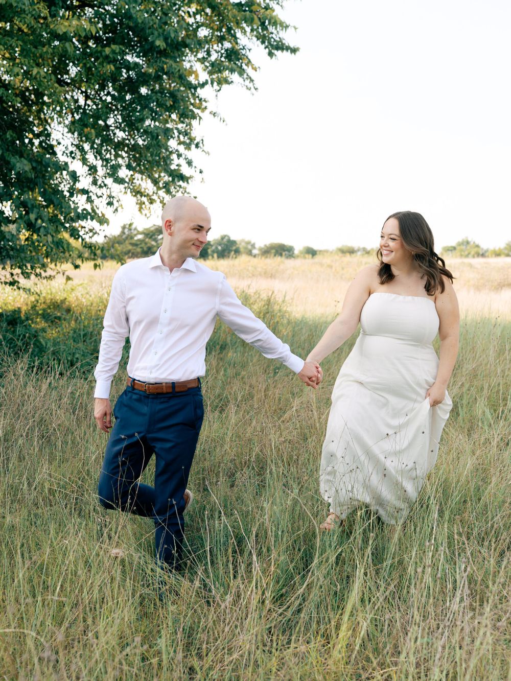 A couple holding hands and walking through tall grass in a field at sunset, with green trees in the background.