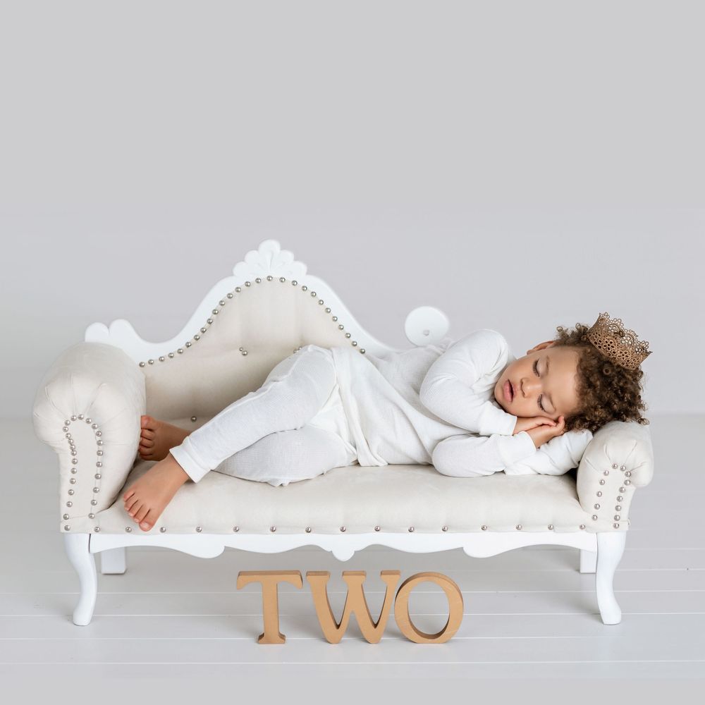 A toddler in white sleeps peacefully on an elegant white tufted chaise lounge with wooden 'TWO' lettering on the floor.