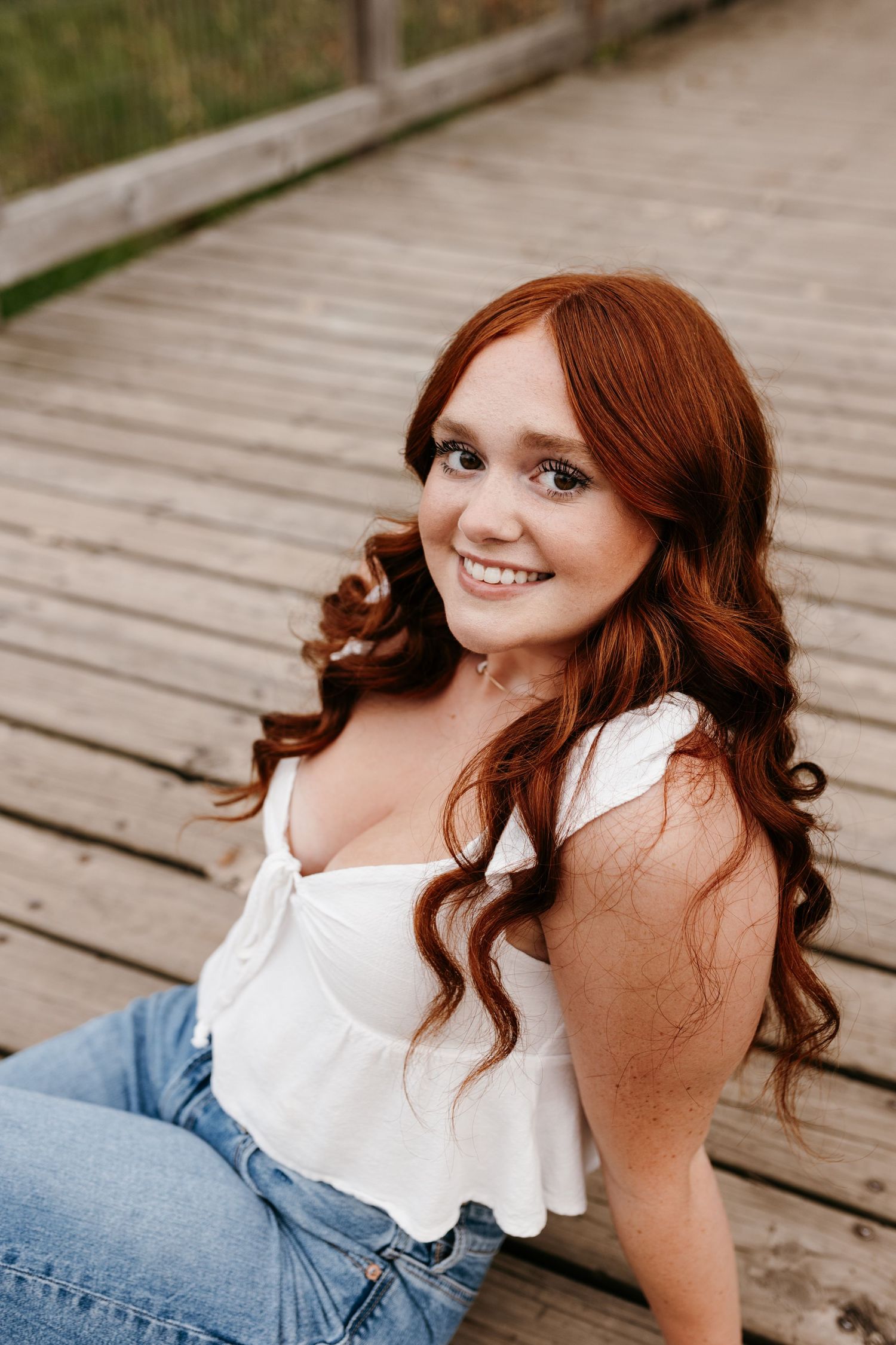 A portrait of someone in a white ruffled top and jeans sitting on a wooden boardwalk.