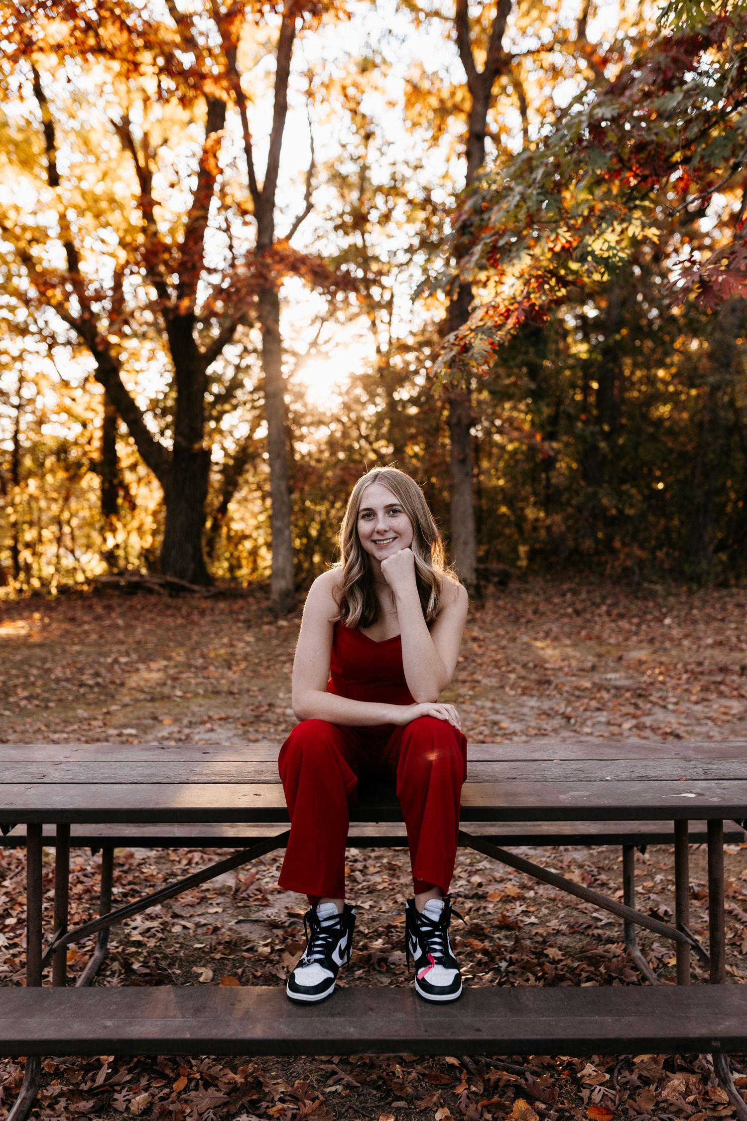Someone in a red outfit sits contemplatively on a picnic table surrounded by fall foliage.