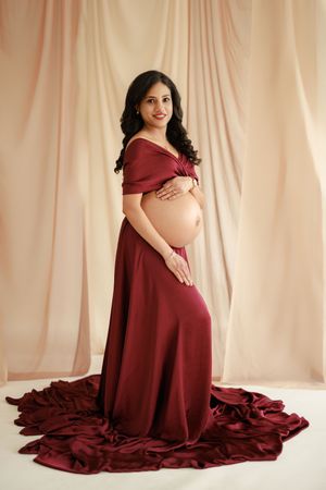 A person in a burgundy flowing maternity gown poses elegantly against a beige backdrop for a pregnancy photo series.