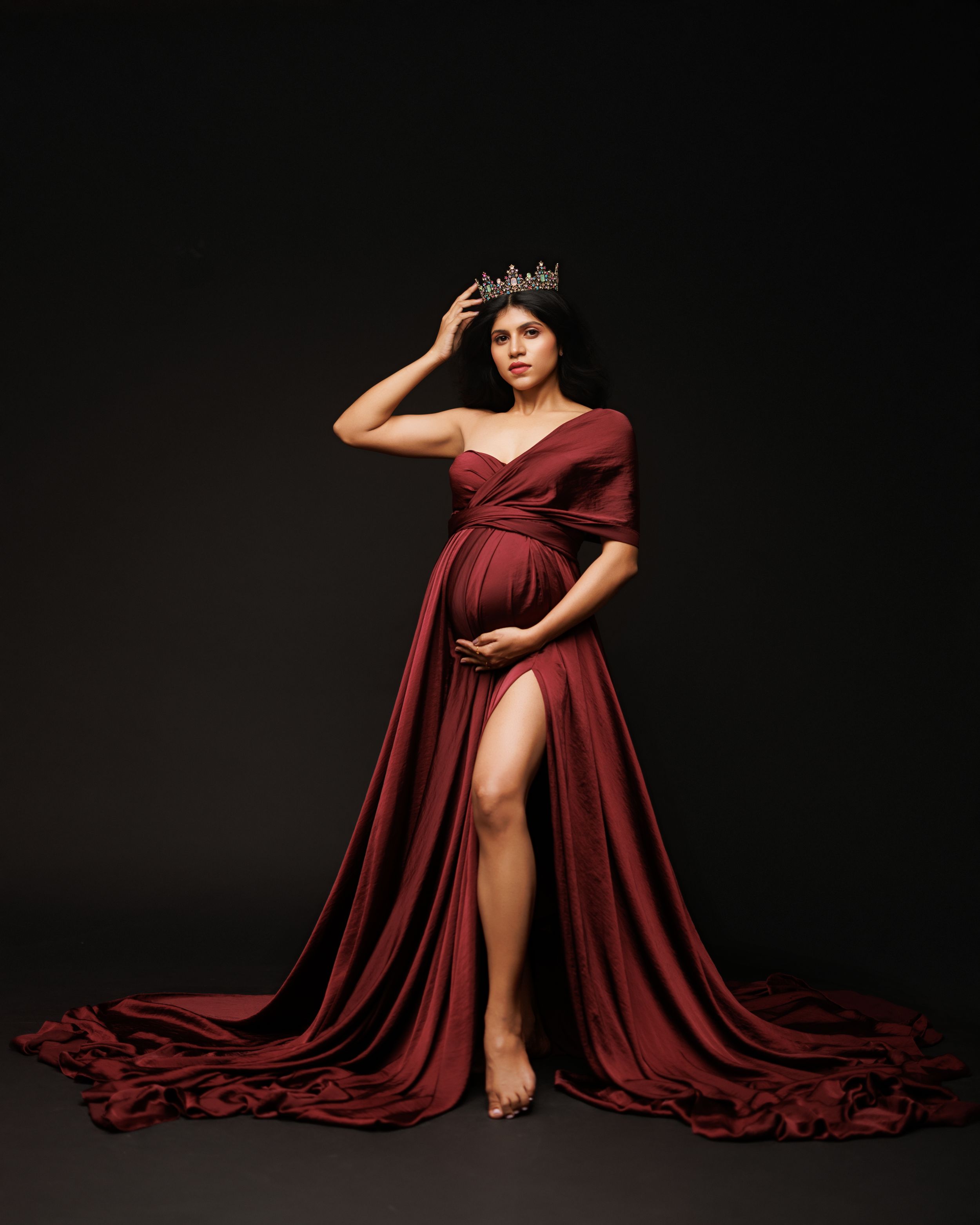 Maternity photo of woman in flowing burgundy gown with train and crown against dark background.