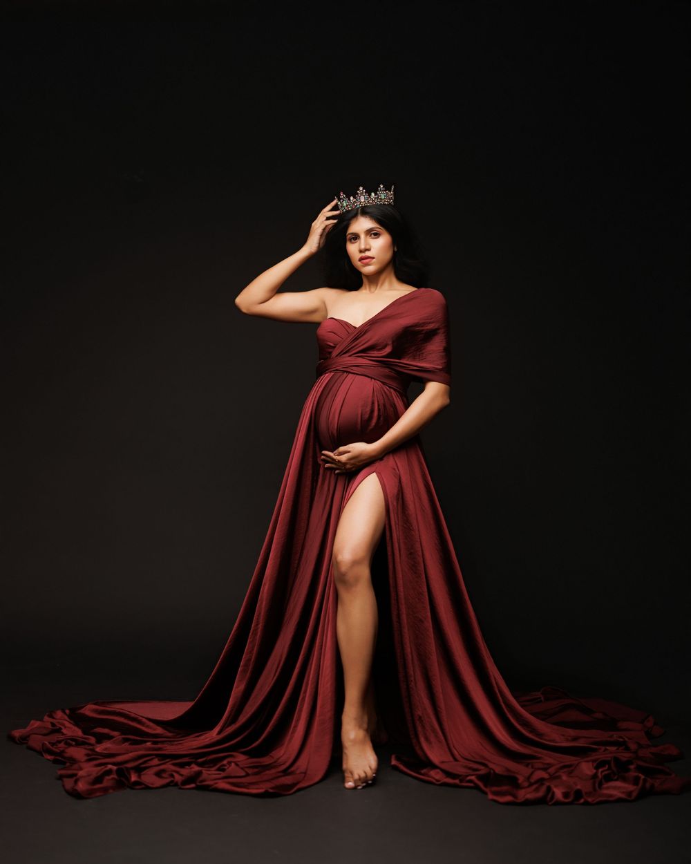 Maternity photo of woman in flowing burgundy gown with train and crown against dark background.