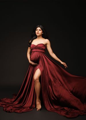 Elegant maternity photo of figure in flowing burgundy gown with dramatic train against dark background.
