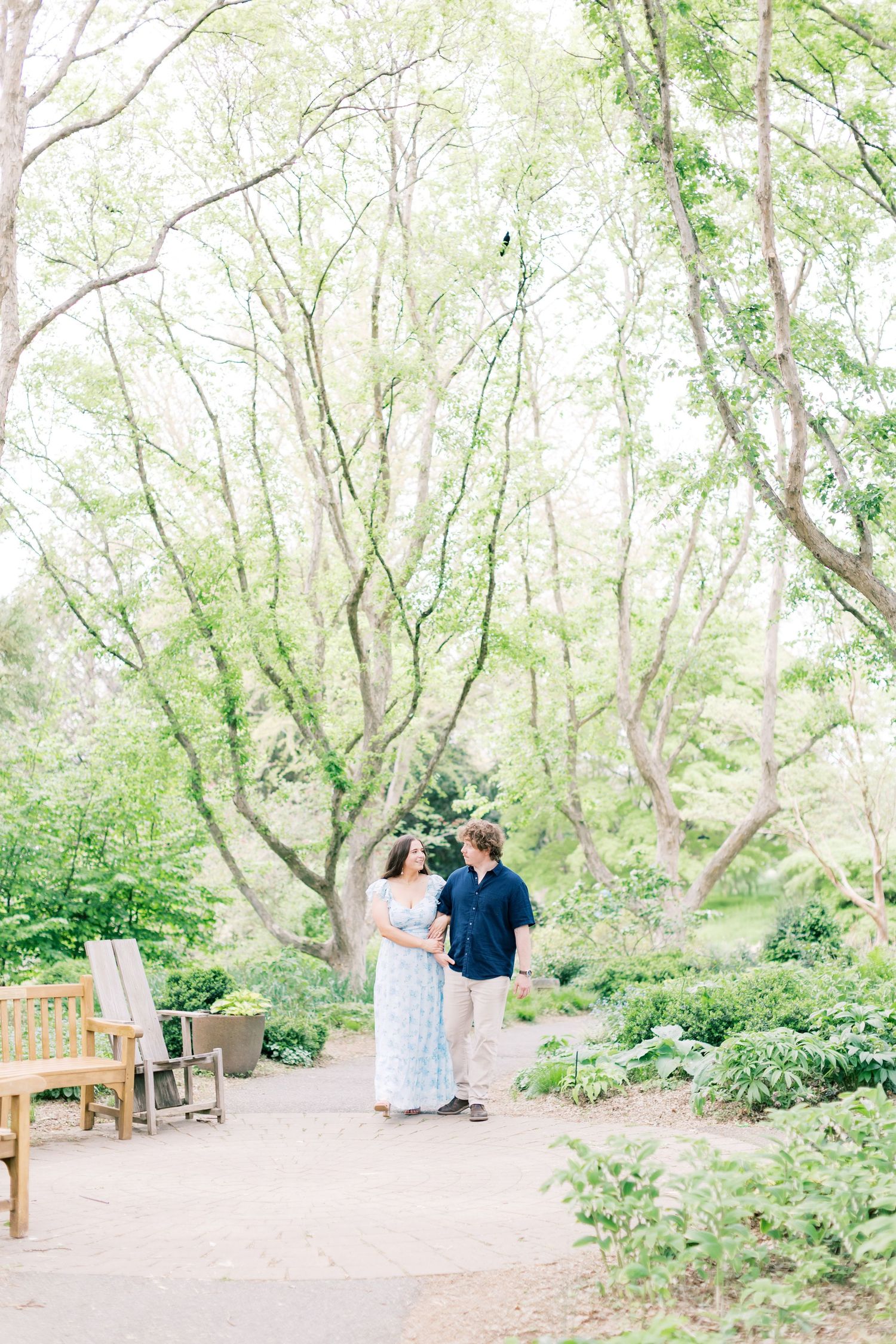 Couple walks hand-in-hand through a lush green garden with towering trees and a wooden bench nearby.