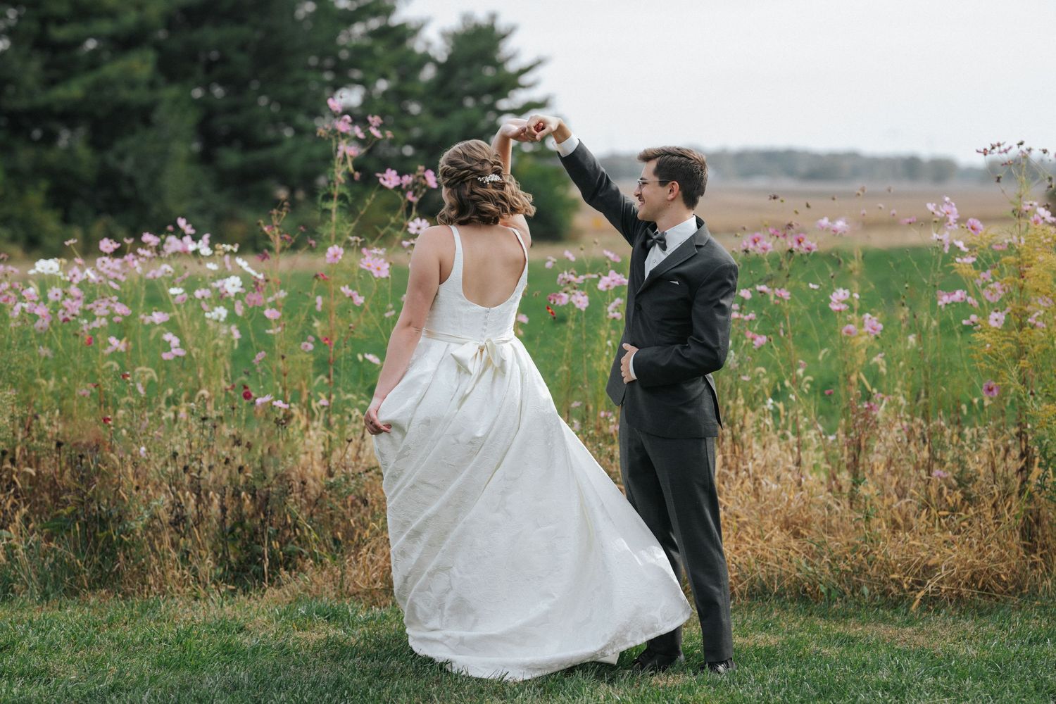 A couple dances together in a wildflower field wearing formal wedding attire on a sunny day.