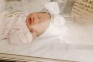 A sleeping newborn baby in white clothing and hat rests against crisp white bedding.
