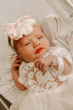 A baby in white lace dress and floral headband poses for a portrait.