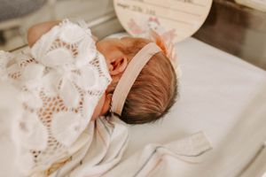 A close-up portrait of a baby wearing a delicate white lace dress and pink headband.