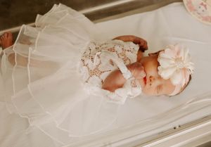 Newborn baby wearing white lace christening gown and bow headband lying on white fabric.