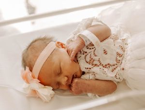 Sleeping newborn in delicate white lace dress with pink bow headband on soft bedding.