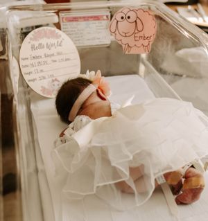 Newborn baby in white dress sleeping in hospital bassinet with decorative wall cards.