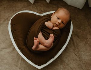 Newborn baby wrapped in brown fabric lying in heart-shaped cushion on beige background.
