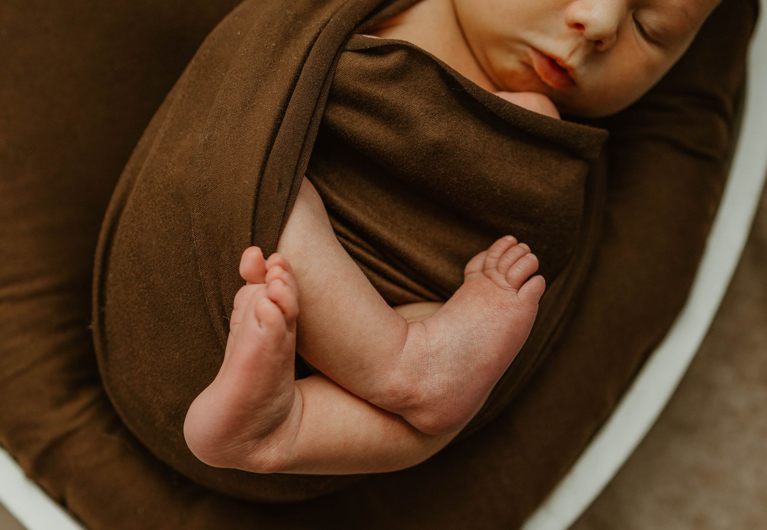 Tiny bare feet of a sleeping baby rest comfortably against soft brown fabric.