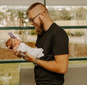 Bearded father in black shirt holding and gazing at his newborn baby with purple bow.