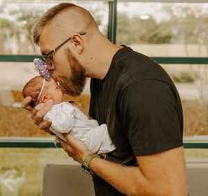 Father in black t-shirt tenderly kissing his newborn baby wearing floral headband.